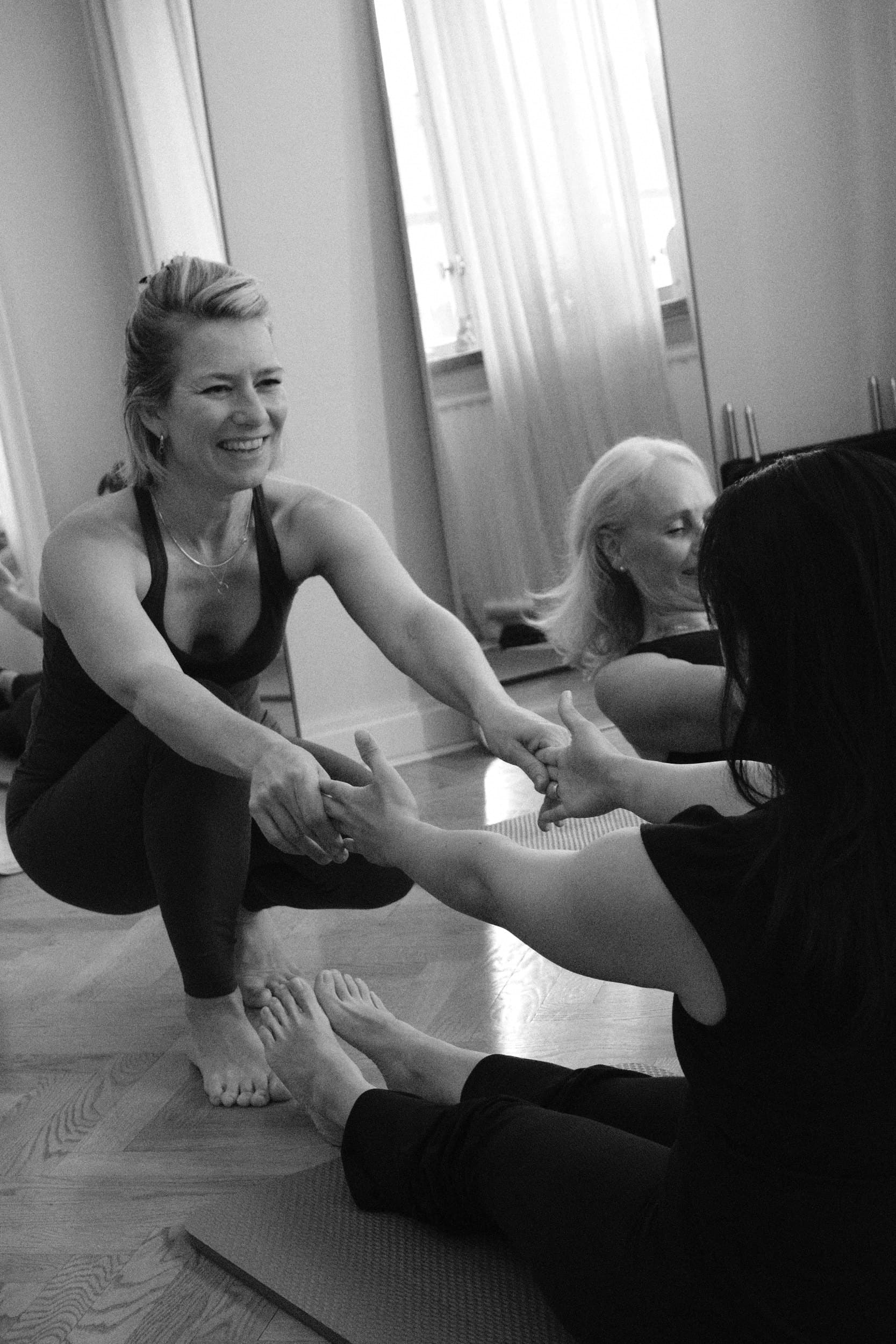 Black and white: Liv, laughing, crouched on her mat and holding hands with a seated client for a partner stretch, another client beside them.