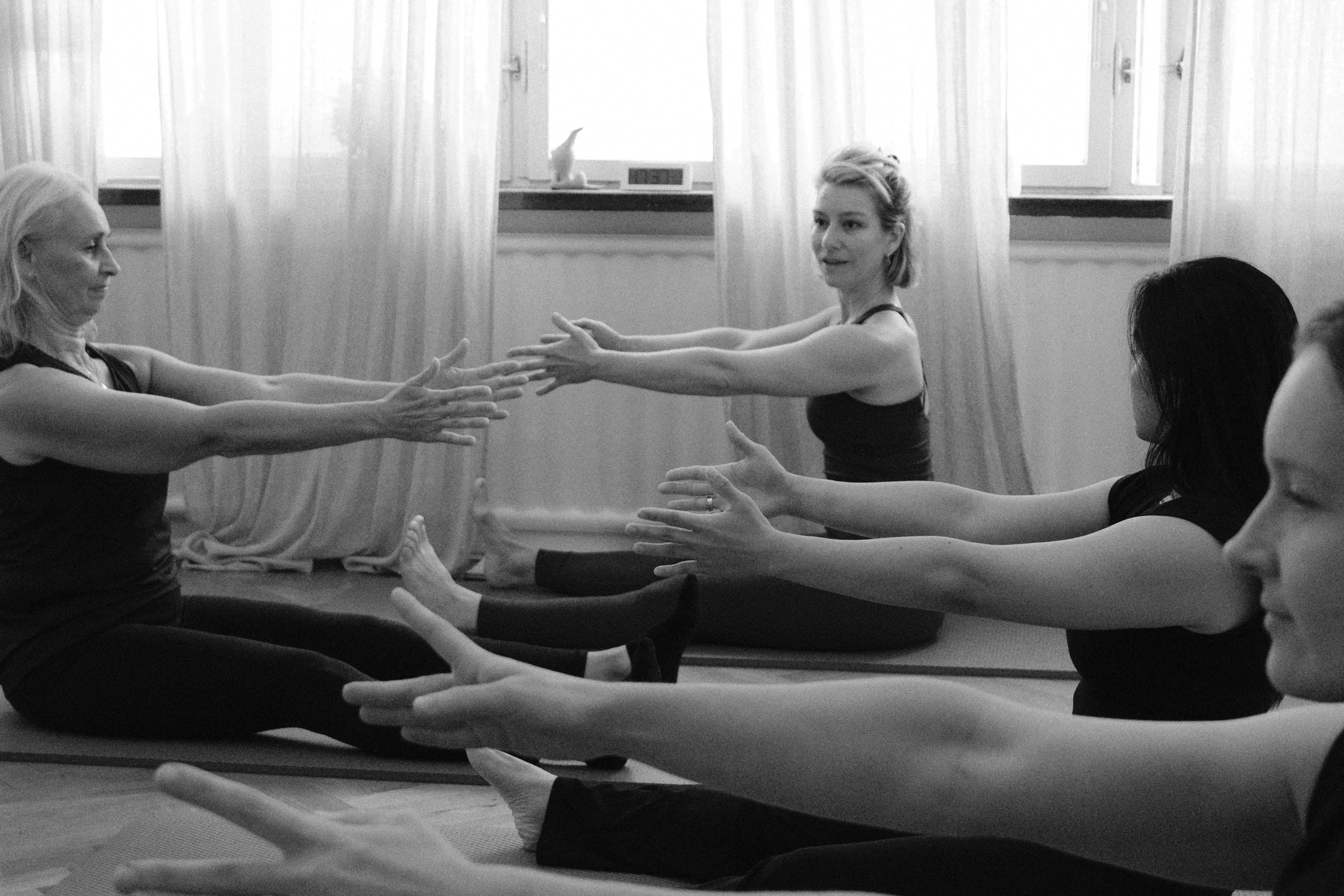 Black and white: a small class sitting on their mats with arms reaching forward, Liv smiling across at them.